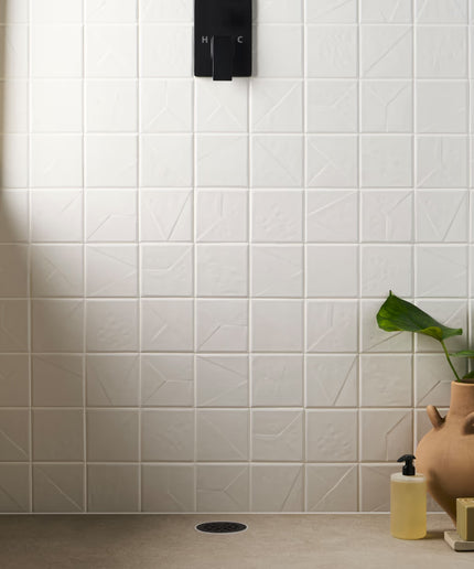 Bathroom corner with white tiled wall, plant, and soap dispenser.