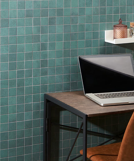 Laptop on a wooden desk against a teal tiled wall with a shelf holding decorative items.