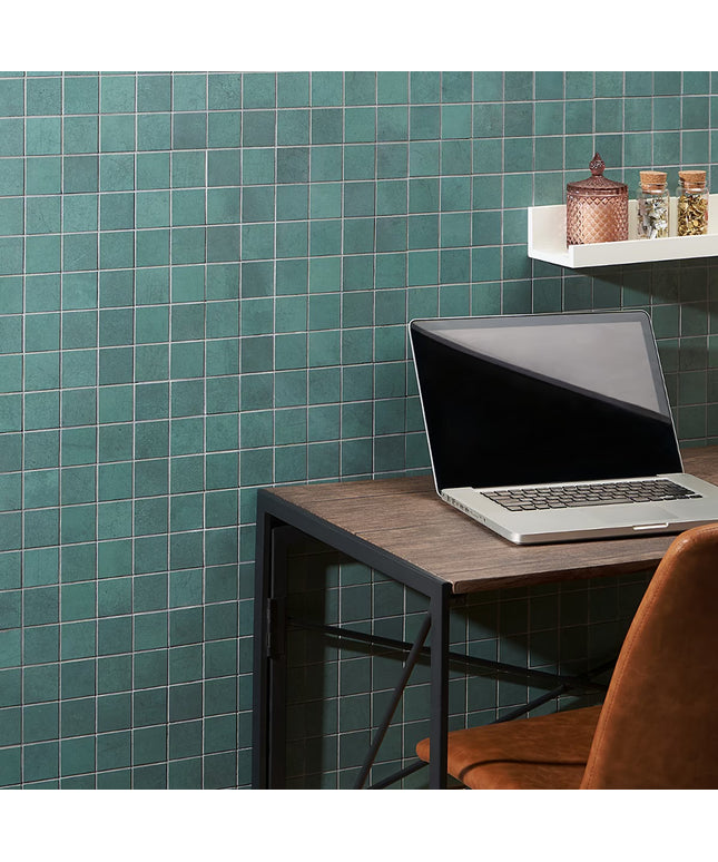 Laptop on a wooden desk against a teal tiled wall with a shelf holding decorative items.