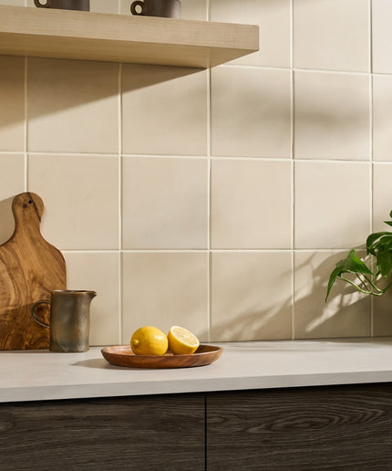 Kitchen counter with wooden cutting board, lemons, and a plant against a tiled wall.