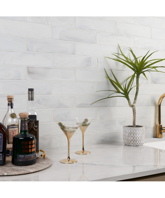Bottles of alcohol, two cocktail glasses, and a potted plant on a kitchen counter with a tiled wall background.