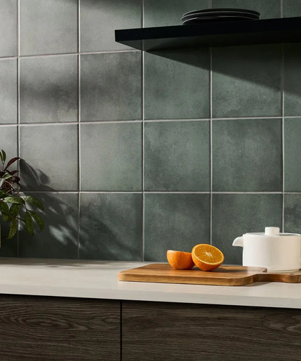 Kitchen counter with oranges on a cutting board and a white teapot against a green tiled wall.
