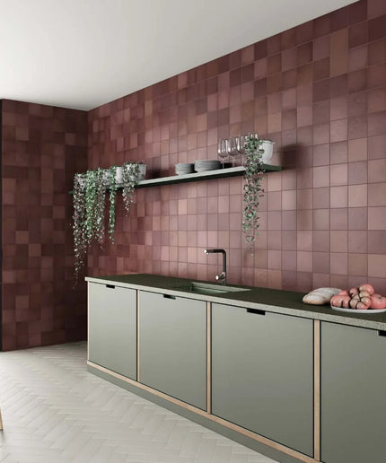 A modern kitchen interior with a wall of square terracotta-brown tiles, sage green cabinets, a dark countertop, and hanging greenery.