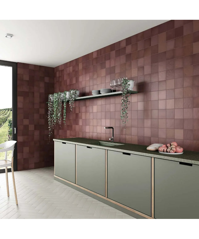 A modern kitchen interior with a wall of square terracotta-brown tiles, sage green cabinets, a dark countertop, and hanging greenery.
