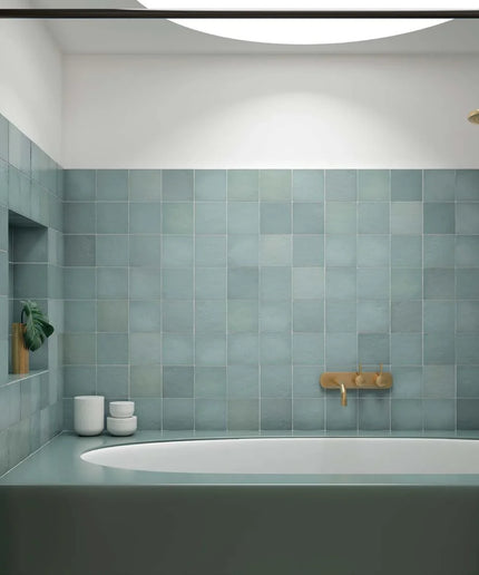 A modern bathroom interior featuring a wall of square matte blue-gray tiles, a matching bathtub, a brass faucet, and a recessed shelf with small white ceramic containers.