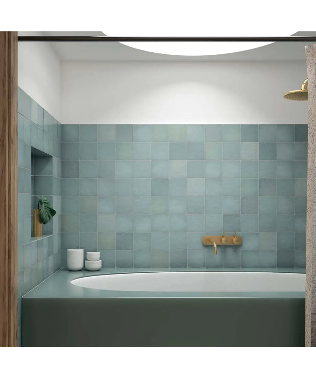 A modern bathroom interior featuring a wall of square matte blue-gray tiles, a matching bathtub, a brass faucet, and a recessed shelf with small white ceramic containers.