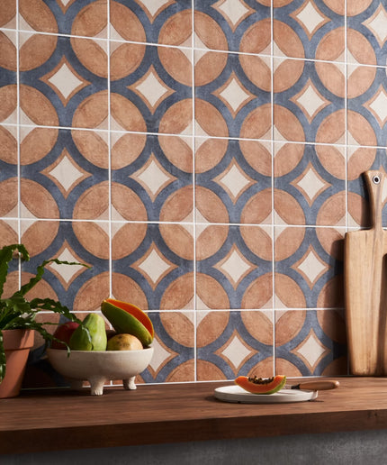 Patterned tiles on a wall with a wooden shelf below containing fruits, a plant, and kitchen utensils.