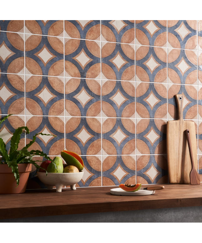 Patterned tiles on a wall with a wooden shelf below containing fruits, a plant, and kitchen utensils.