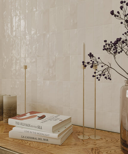 Stack of books on a wooden surface with decorative candles and a vase with branches against a tiled wall.