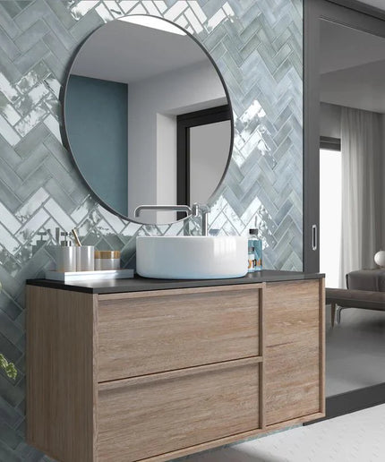 Glossy light blue herringbone wall tiles in modern bathroom with wood vanity and round mirror.