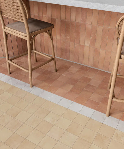 Two wooden bar stools with woven cane backs and brown cushioned seats placed in front of a terracotta tiled wall, with a light stone countertop above and a beige tile floor below.