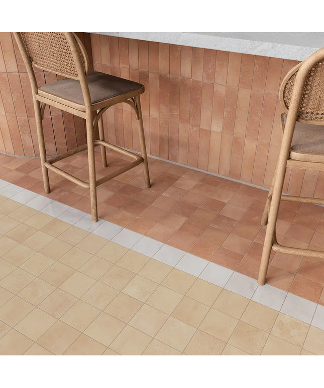 Two wooden bar stools with woven cane backs and brown cushioned seats placed in front of a terracotta tiled wall, with a light stone countertop above and a beige tile floor below.